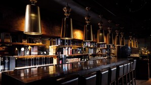 Un bar con varias lámparas colgantes, una barra larga con taburetes y estantes llenos de botellas y vasos.