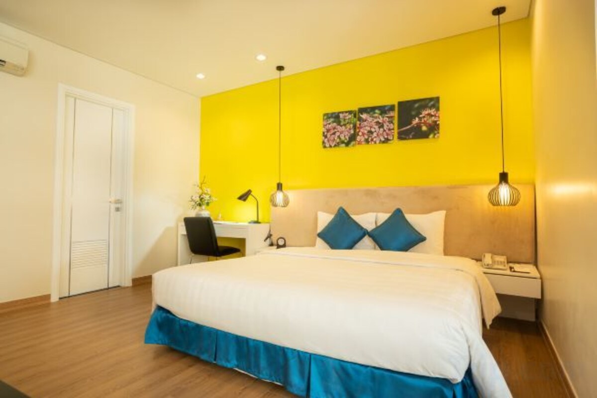 A hotel room with a large bed, a desk, and a chair. There is a door on the left and a nightstand with a lamp on the right.