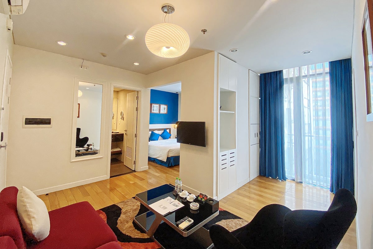 A modern hotel room with a living area, a bedroom with a bed and blue walls, a wardrobe, and a large window with curtains.