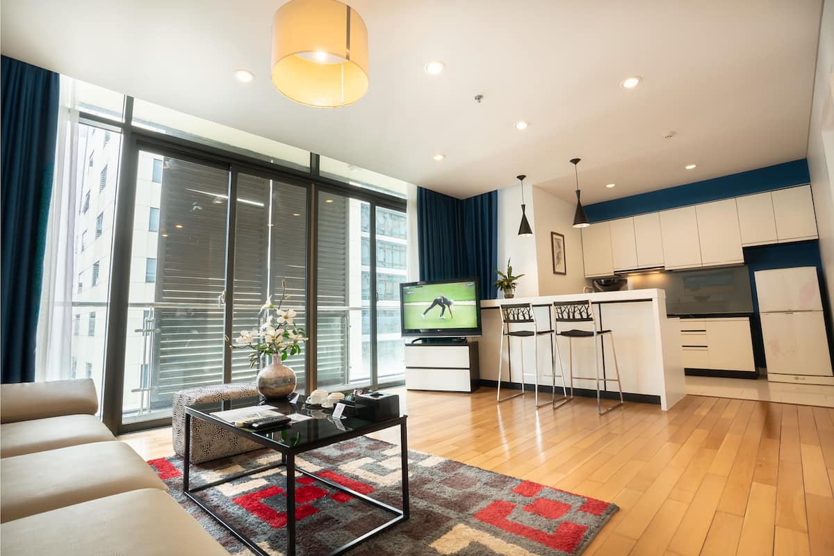 A modern living room with a sofa, a coffee table, and a TV. There is a kitchen area with a fridge, a stove, and a sink. The room has large windows and a view of the city.