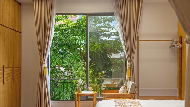 Une chambre avec une grande fenêtre, un lit, une armoire en bois, une petite table avec un vase et une chaise.