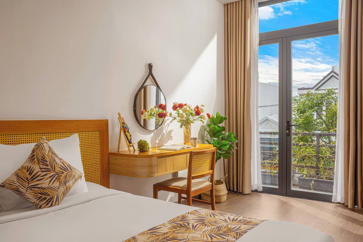 A hotel room with a bed, a desk with a mirror, a vase with flowers, and a view of the outside.