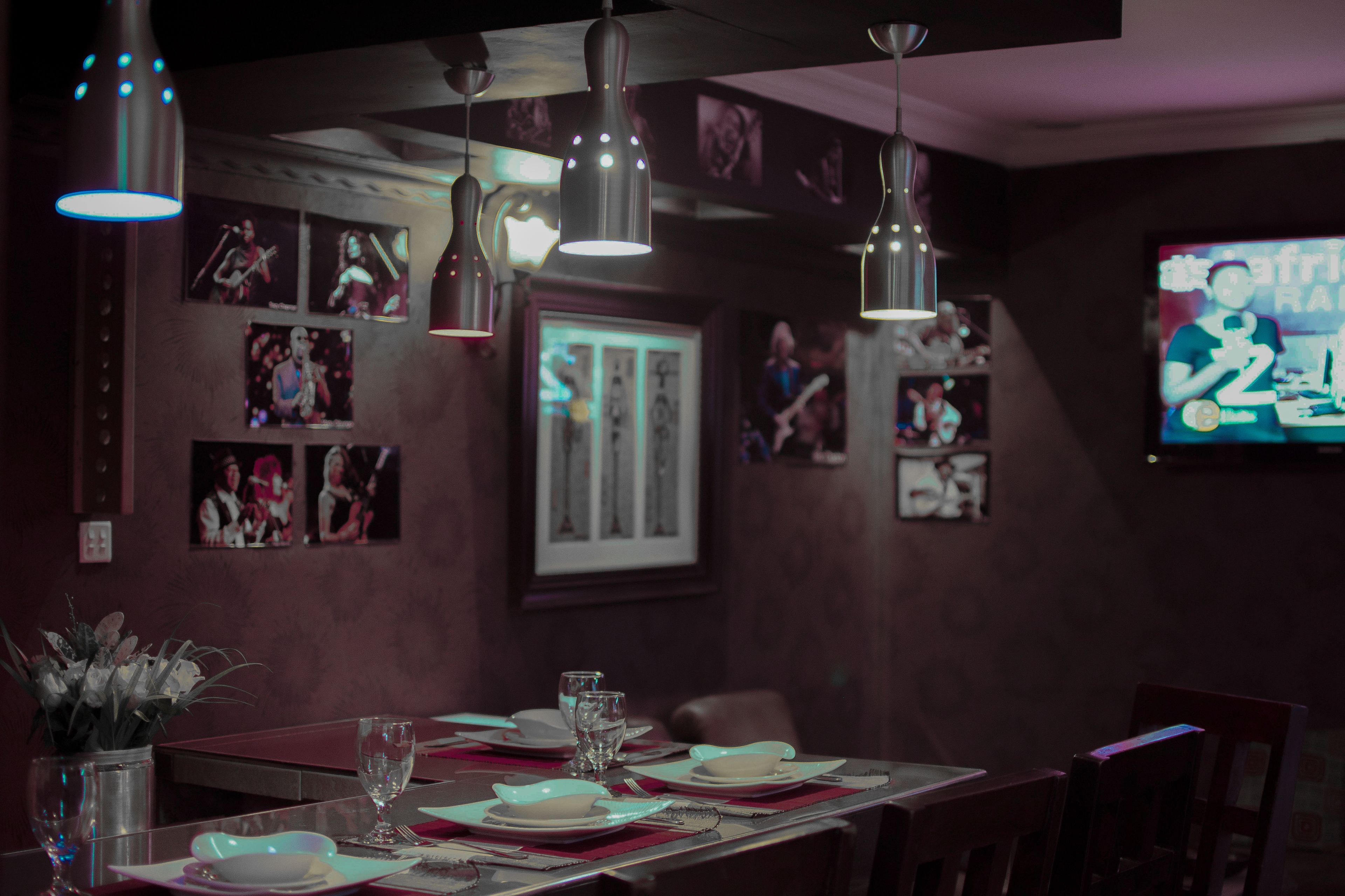 A dining area with a table set for two, overhead pendant lighting, framed pictures on the wall, and a television screen displaying a program.