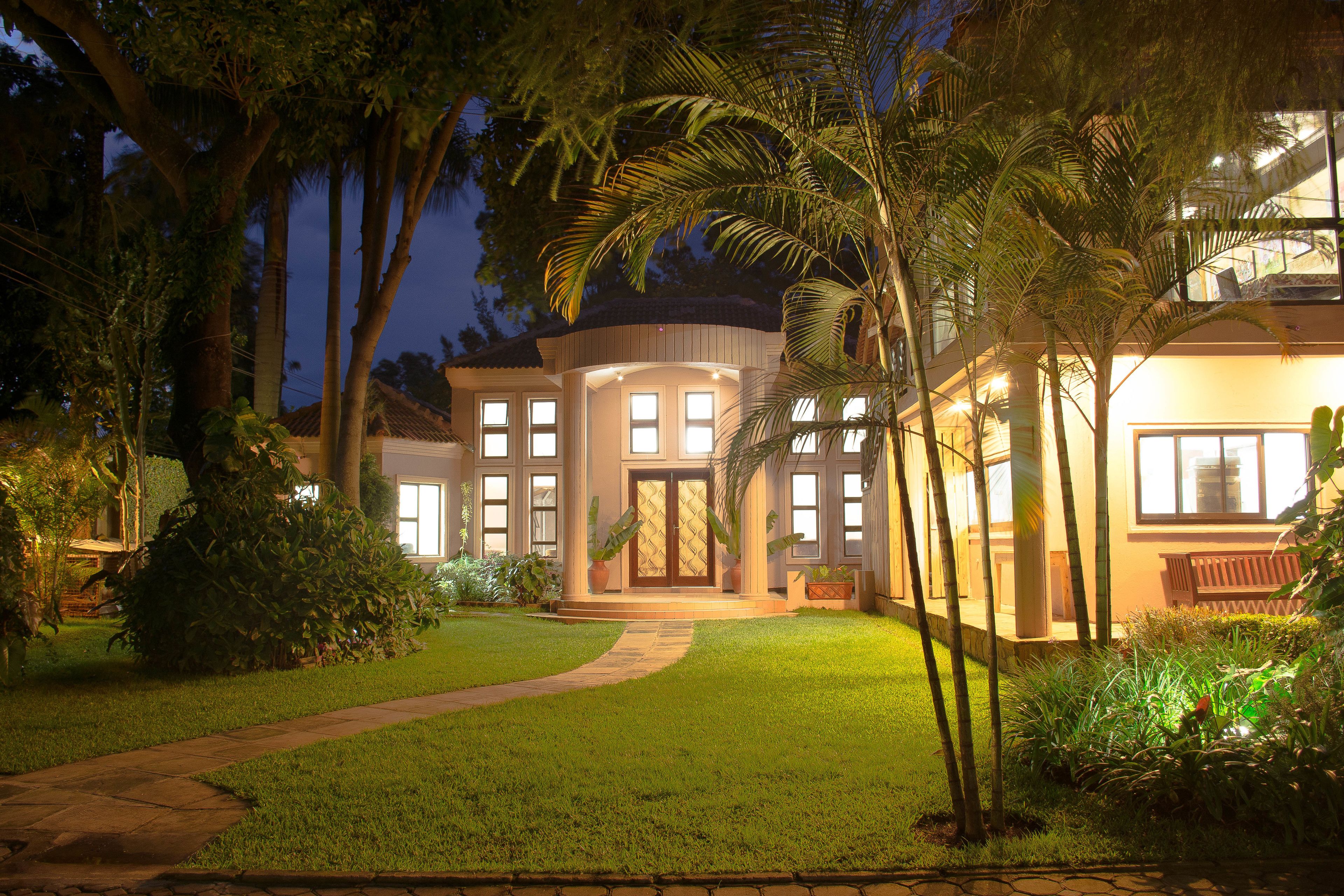A well-lit, two-story house with a curved roof, surrounded by lush greenery and palm trees.