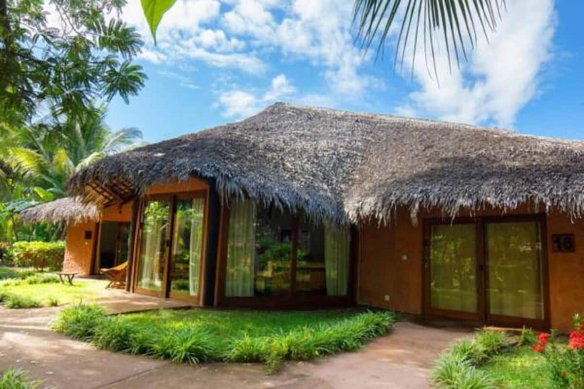 Un bungalow au toit de chaume doté de grandes portes-fenêtres coulissantes, d’une terrasse et d’une végétation luxuriante.