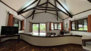 A spacious room with a curved wooden counter, large windows with curtains, a flat-screen TV, and a patterned wooden floor.