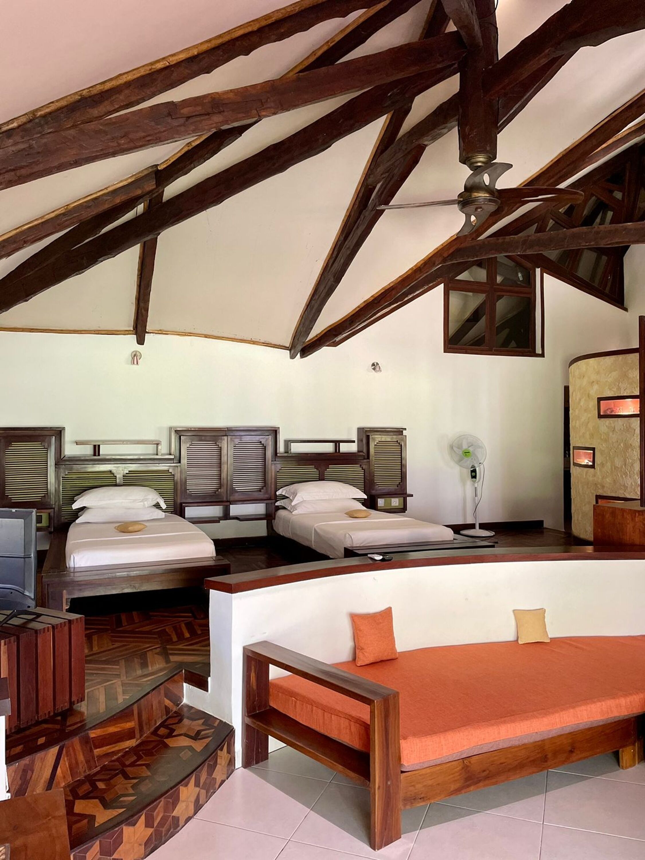 A room with a high ceiling, exposed wooden beams, and a sloped ceiling. There are three beds with wooden headboards, a wooden bench, and a desk with a chair.