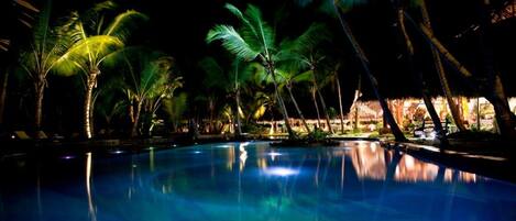 Un área de piscina nocturna con palmeras y una estructura de techo de paja al fondo.