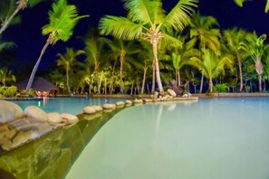 Une zone de piscine éclairée la nuit, avec des palmiers et un petit bâtiment en arrière-plan.