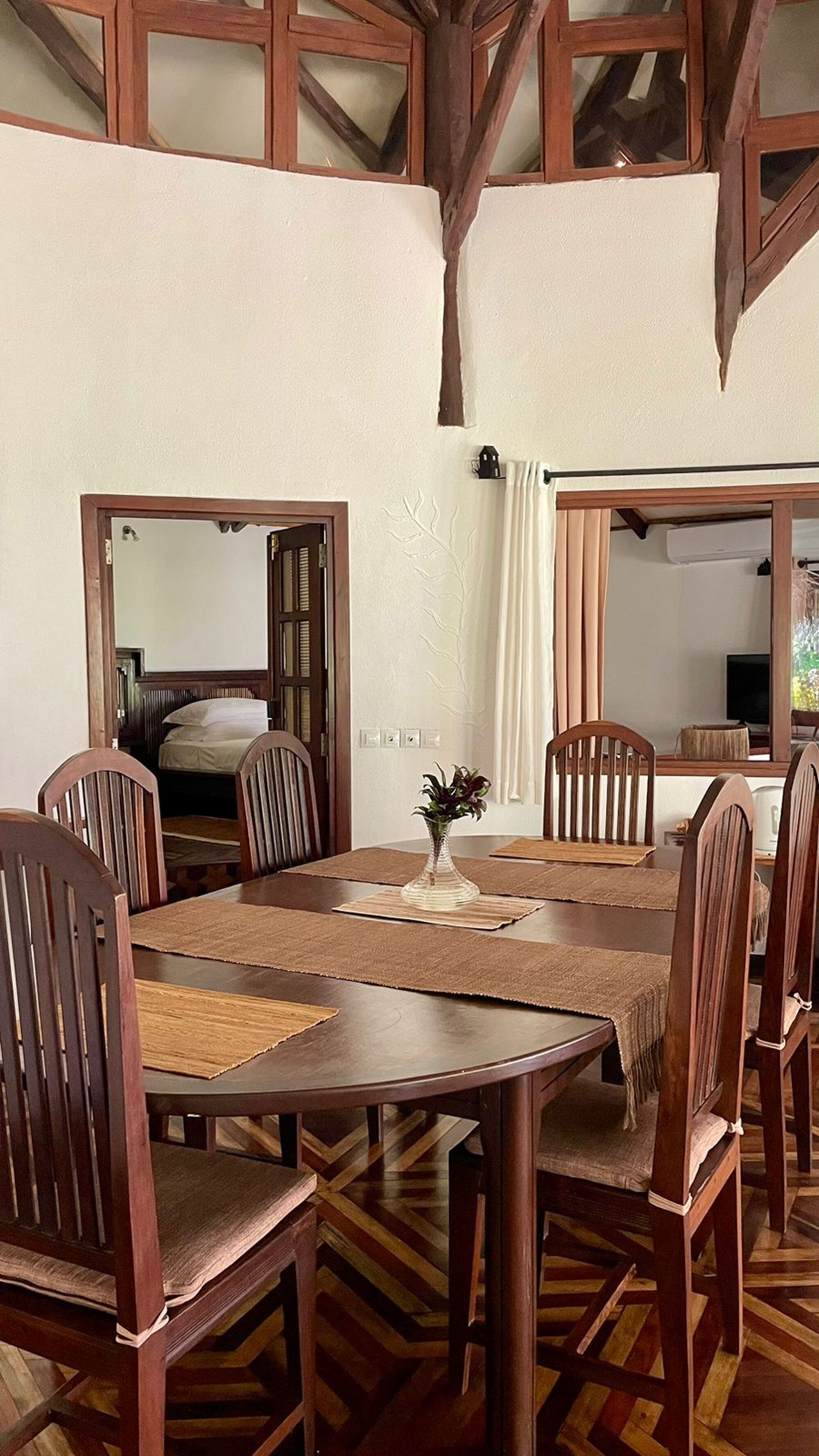 A dining area with a wooden table and chairs, a vase with flowers, and a mirror reflecting the room.