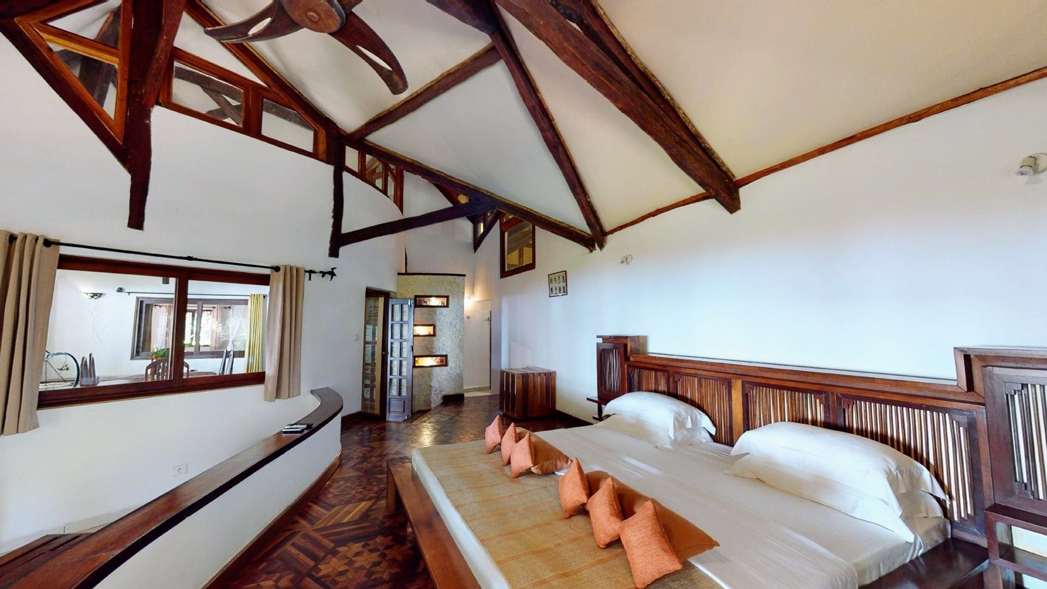Ein Zimmer mit einem großen Bett, Holzbalken, einem Fenster mit Vorhängen und Blick ins Freie.