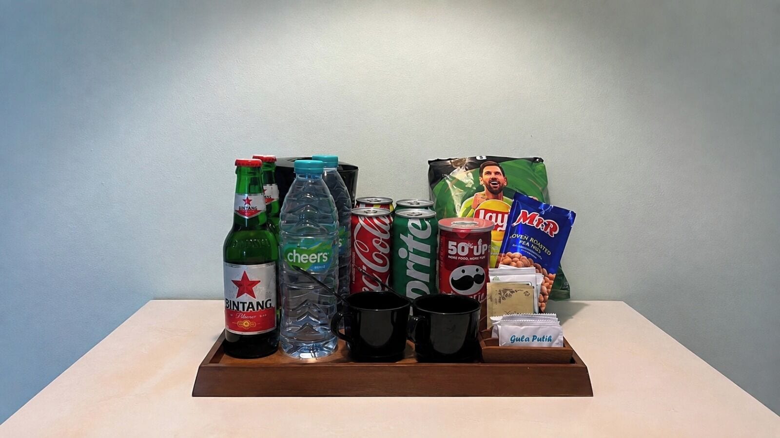 Una bandeja con bebidas y aperitivos, incluyendo una botella de Bintang, una botella de Cheers, una lata de Sprite, una lata de Coca-Cola, una bolsa de MMR y un paquete de Gula Putih.