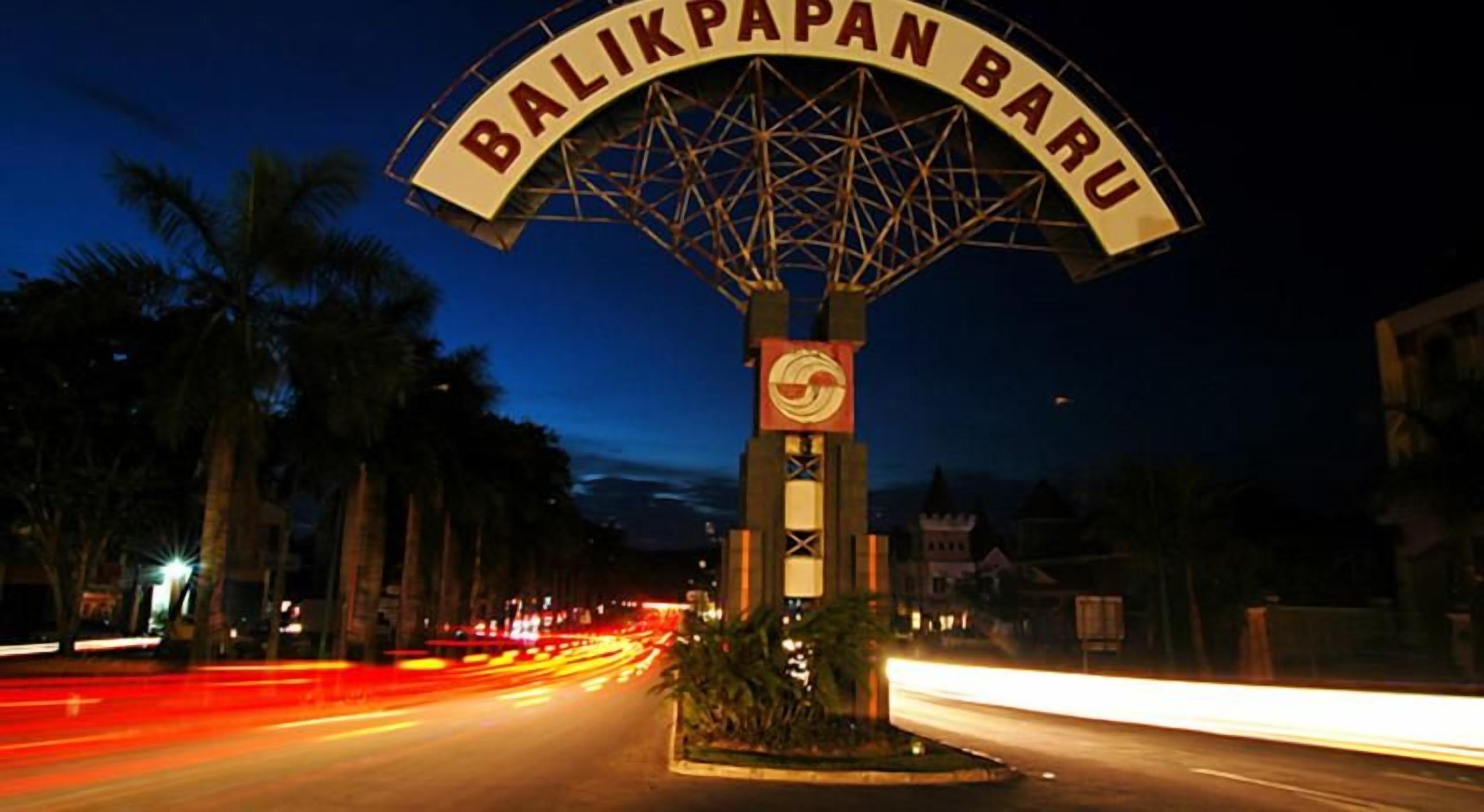 Vista nocturna de una calle con un gran cartel que dice 'BALIKPAPAN BARU' y una torre del reloj.