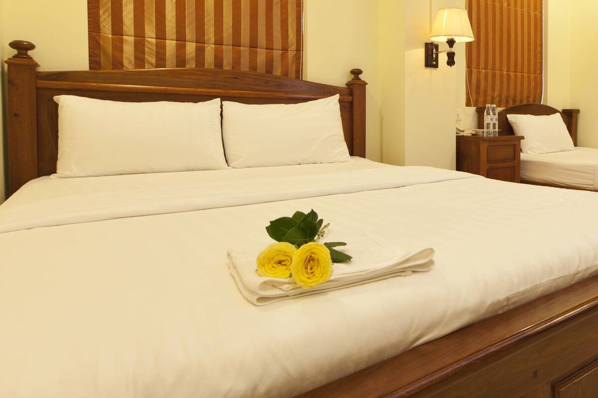 Ein Hotelzimmer mit einem großen Bett, einem kleineren Bett, Holz-Kopfteilen, einem Nachttisch mit Lampe und einem Fenster mit gestreifter Gardine.