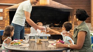 En familie spiser udendørs på en restaurant med en murstensovn og et træbord.