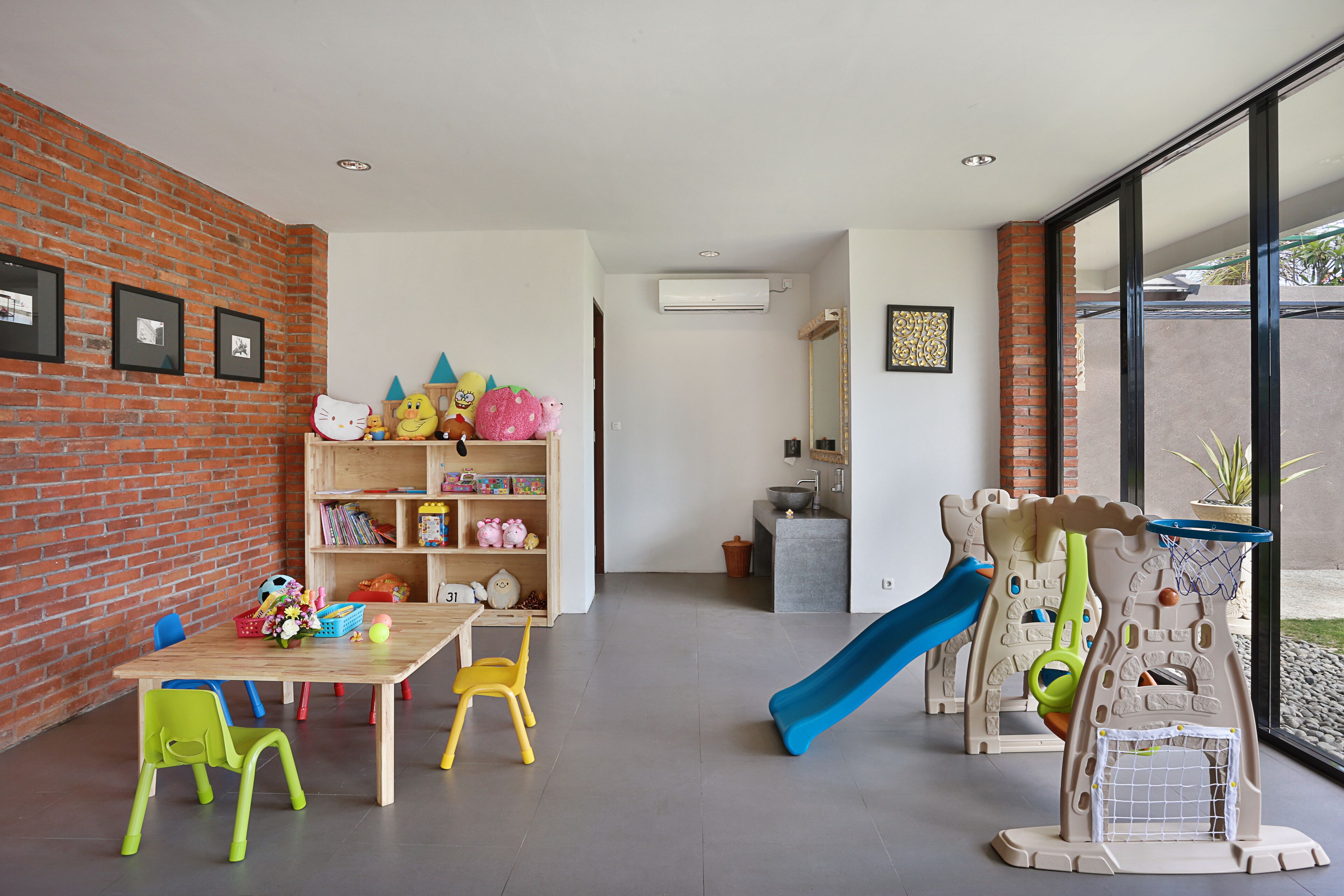 A room with a brick wall, a bookshelf, a table with chairs, a slide, and a play area.