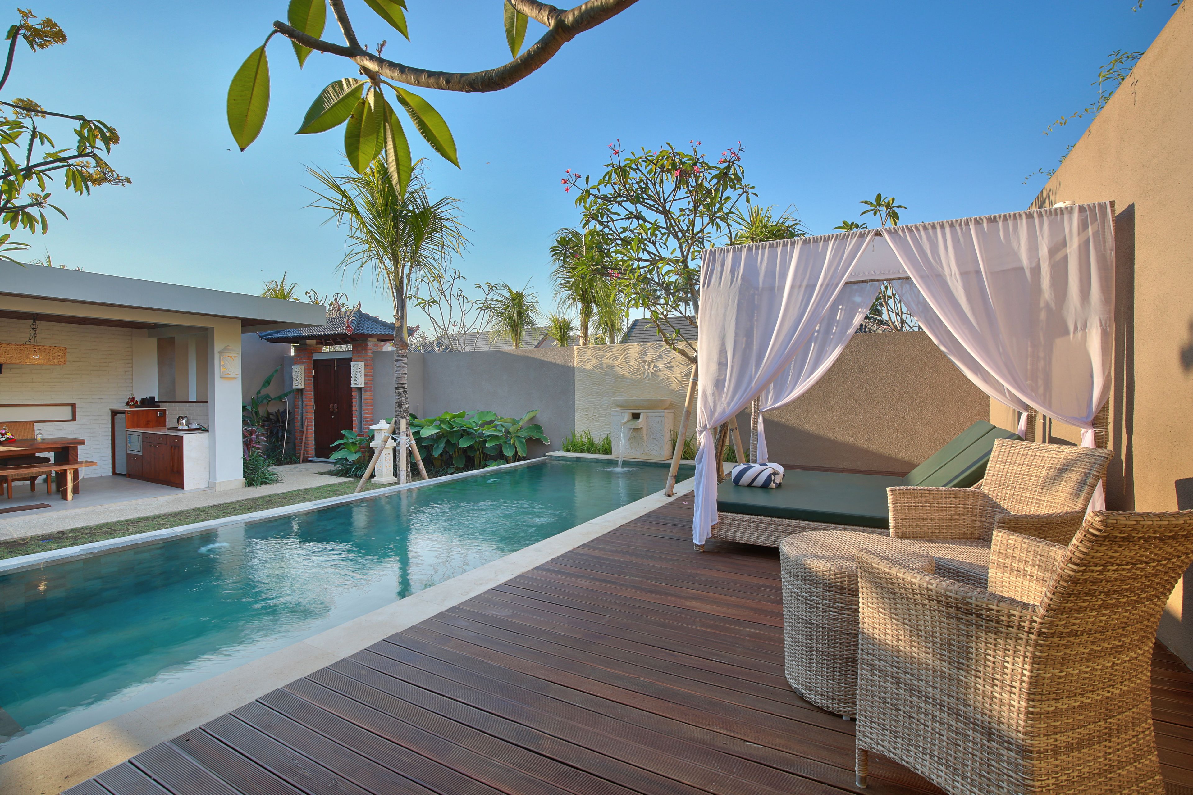 Un área junto a la piscina con un deck de madera, un sofá de mimbre con dosel y una pequeña cocina.