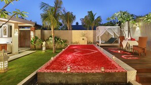 Ein Pool mit roten Blütenblättern, ein Essbereich mit Stühlen und ein Gebäude mit einem Balkon.