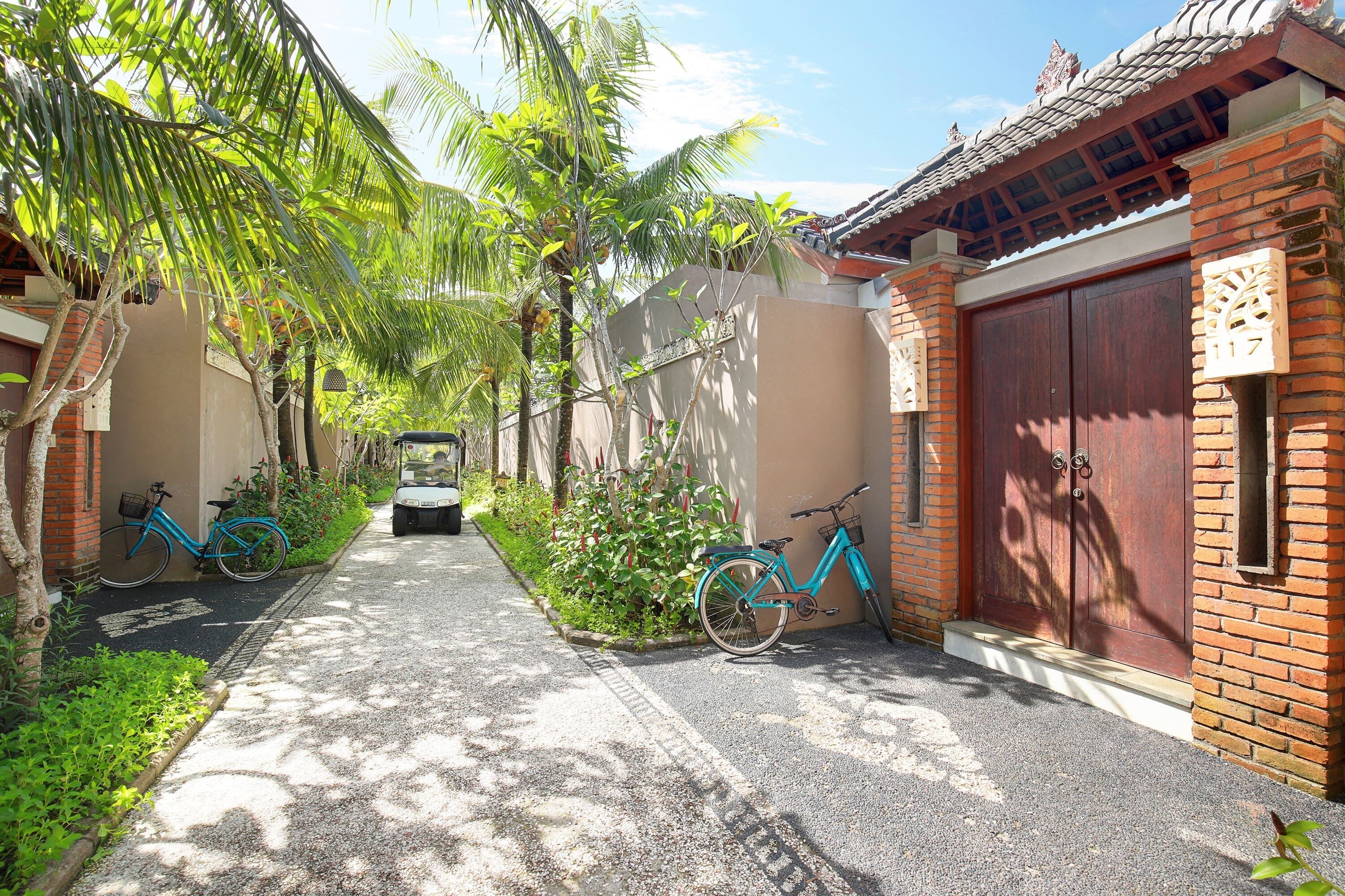 A narrow pathway flanked by lush greenery and a traditional building with a tiled roof and a large wooden door.