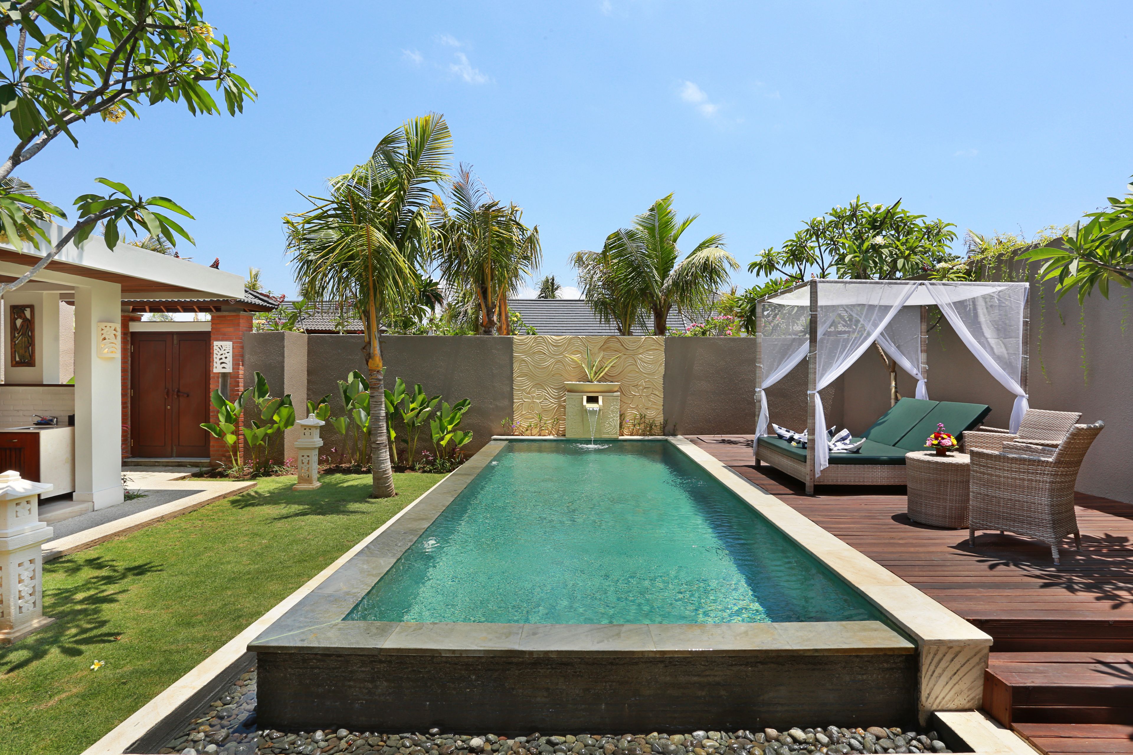 A rectangular swimming pool with a stone border, surrounded by a wooden deck, palm trees, and a small waterfall feature.
