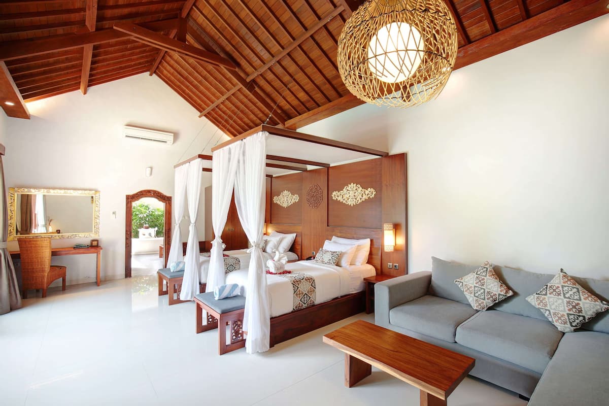 Een bed met baldakijn met wit beddengoed, een houten bankje, een grijze bank met gekleurde kussens en een houten bijzettafeltje in een goed verlichte kamer met een hoge nok en een geweven lamp.