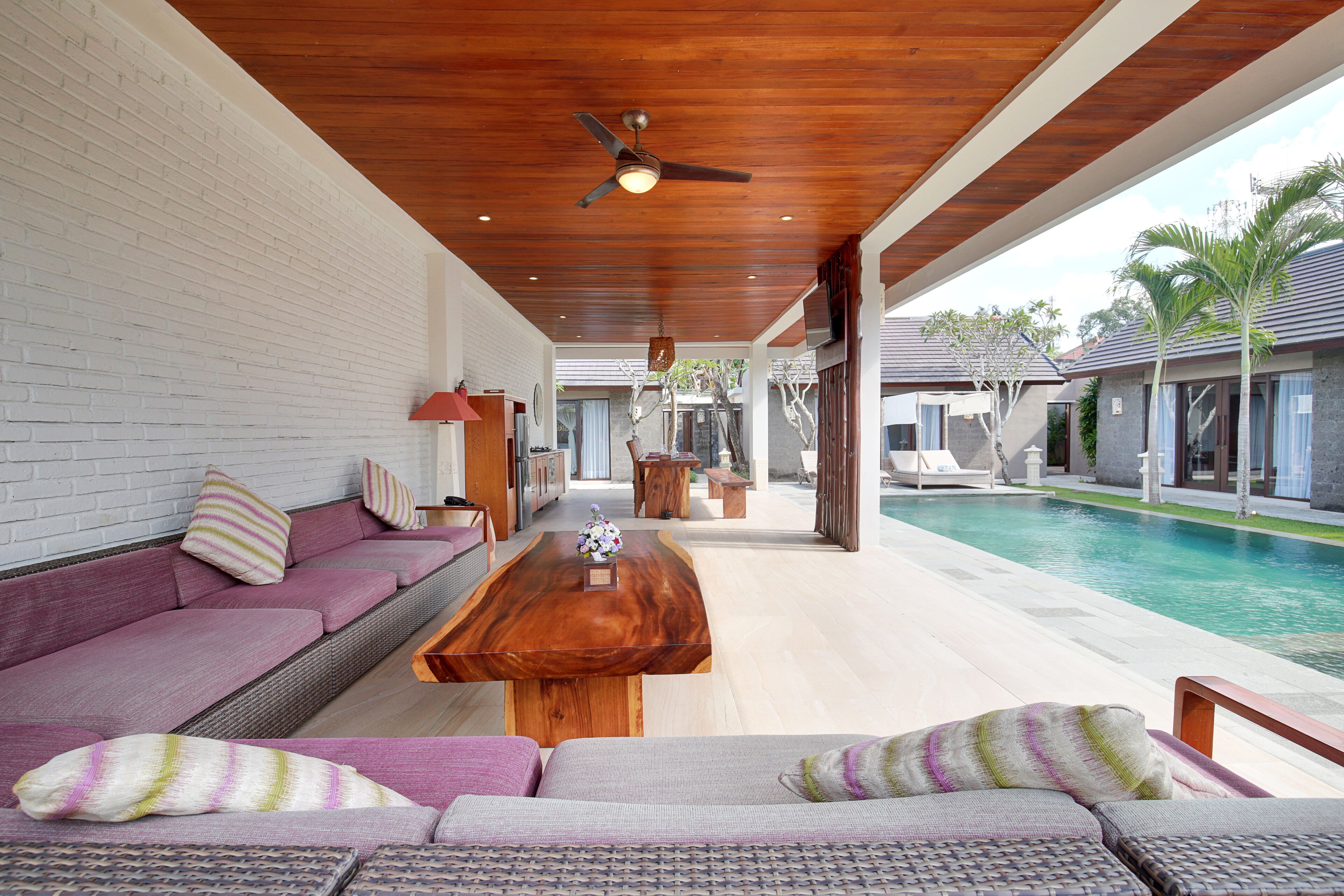 A poolside area with a wooden table, purple and gray sofas, and a ceiling fan.