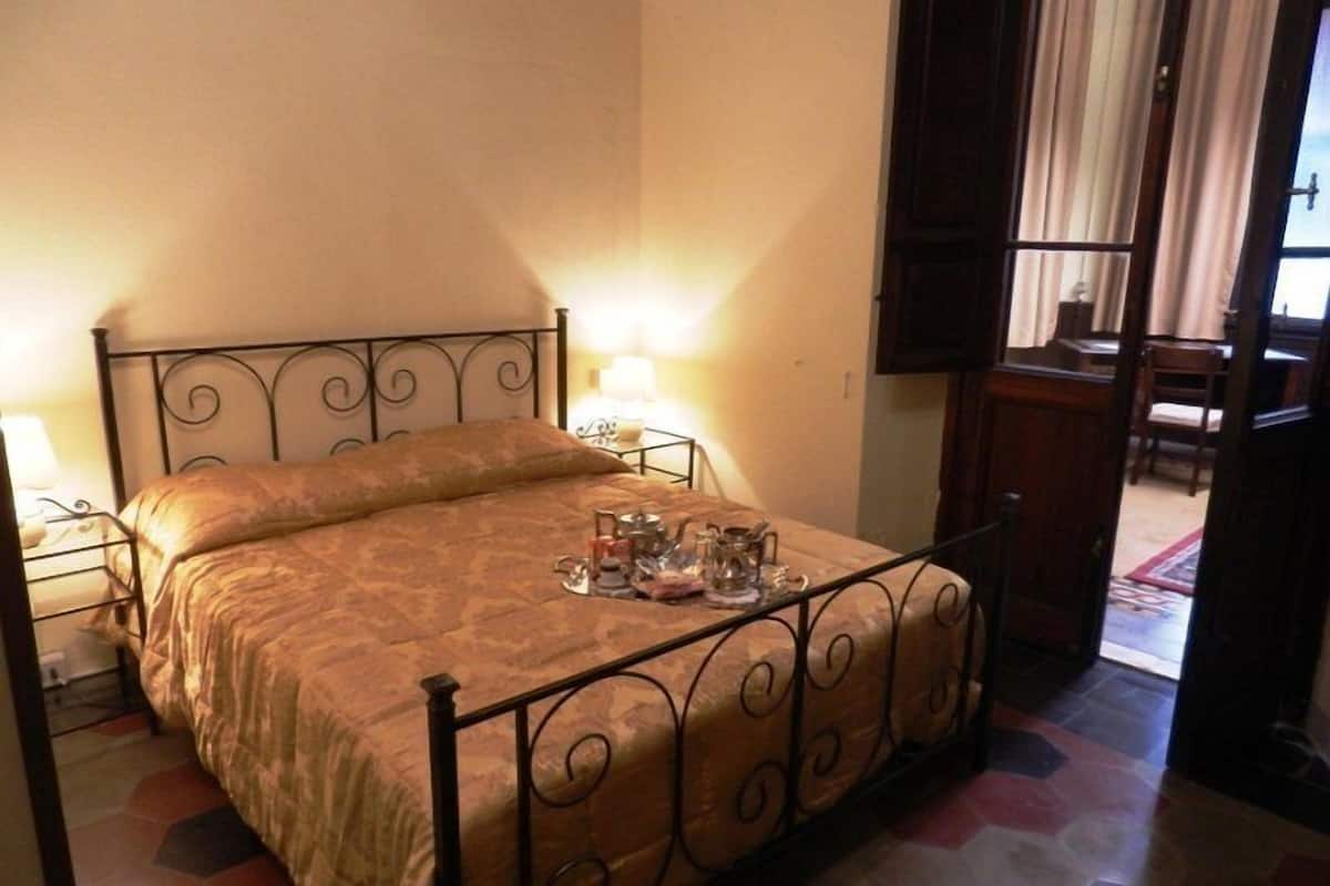 Ein Einzelbett mit Metallgestell und einem gemusterten Bettbezug, ein Nachttisch mit Lampe sowie Blick auf einen Essbereich durch eine offene Tür.