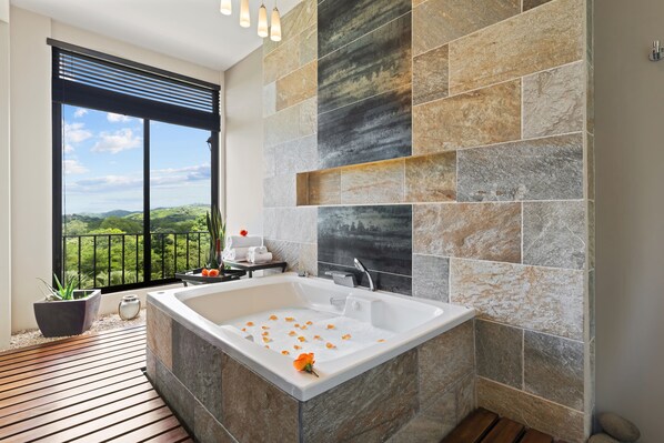 A modern bathroom with a large bathtub, a view of greenery through a sliding door, and a stone-tiled wall.