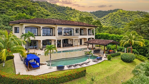Una casa de dos pisos con piscina, rodeada de vegetación exuberante y colinas.