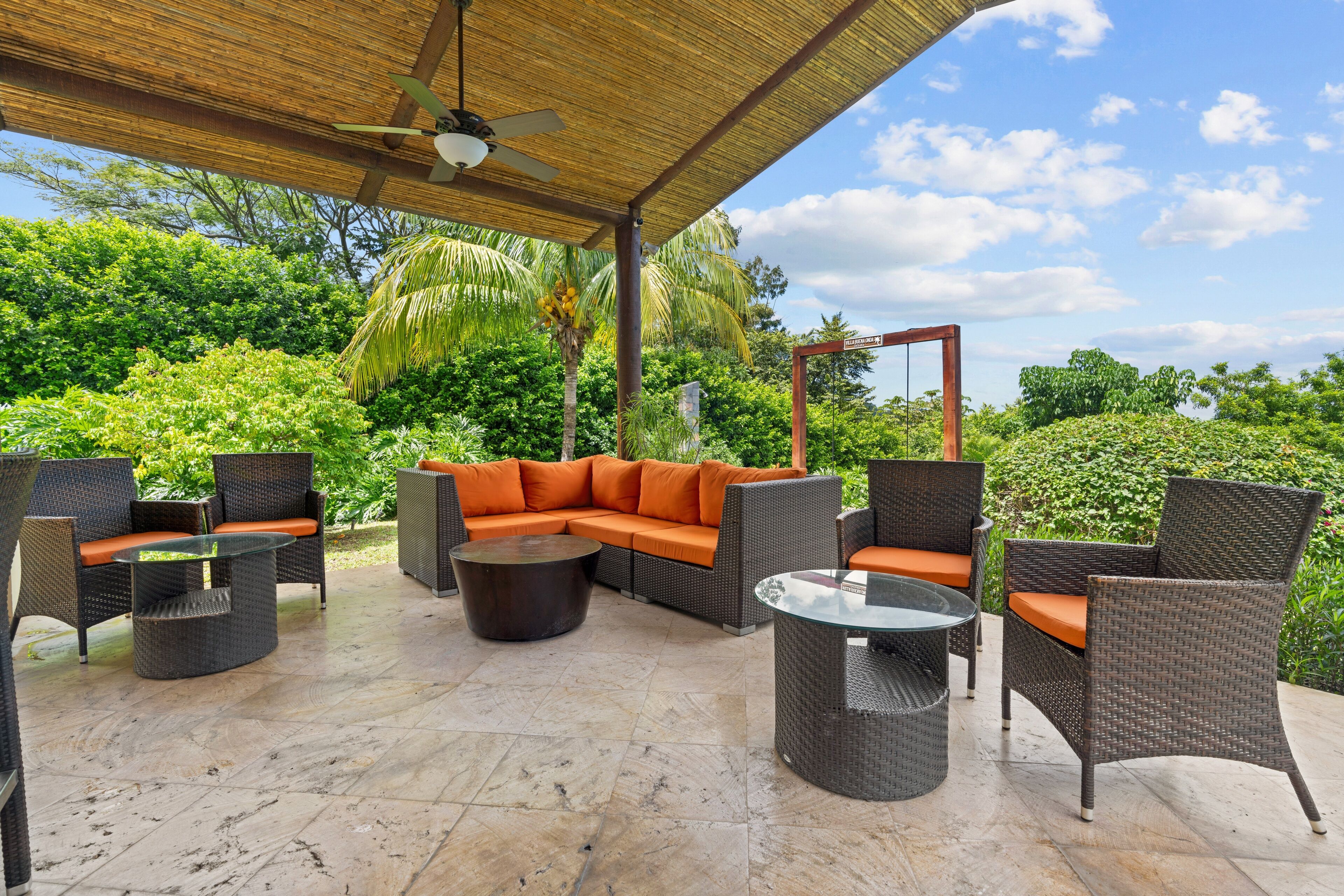 Un patio exterior con mobiliario de mimbre, una mesa de vidrio y un ventilador de techo.