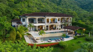 Una amplia casa de dos pisos con piscina, mobiliario exterior y exuberante vegetación.