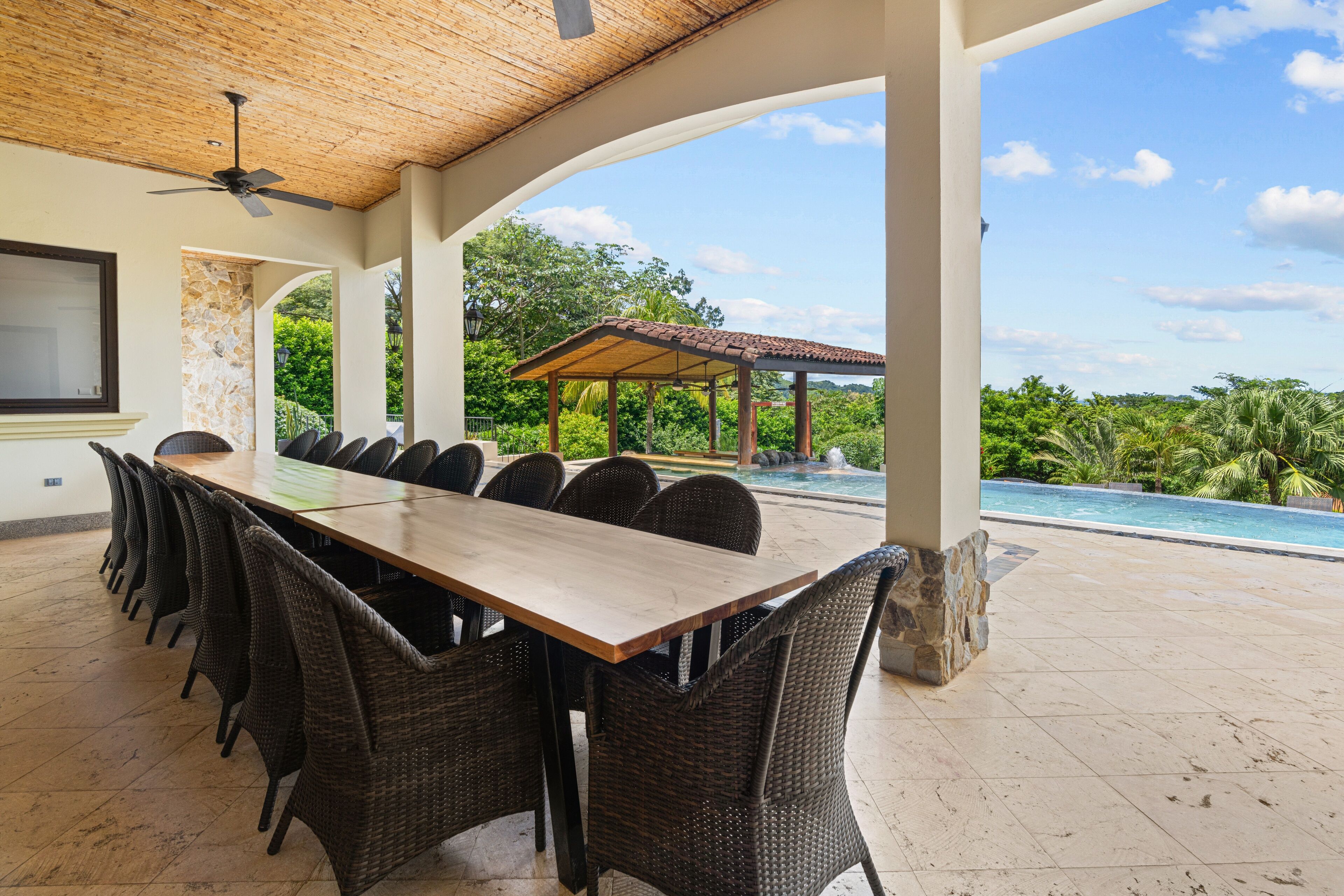Uma área de refeições coberta ao ar livre, com uma mesa comprida e cadeiras de vime, com vista para uma piscina e vegetação exuberante.