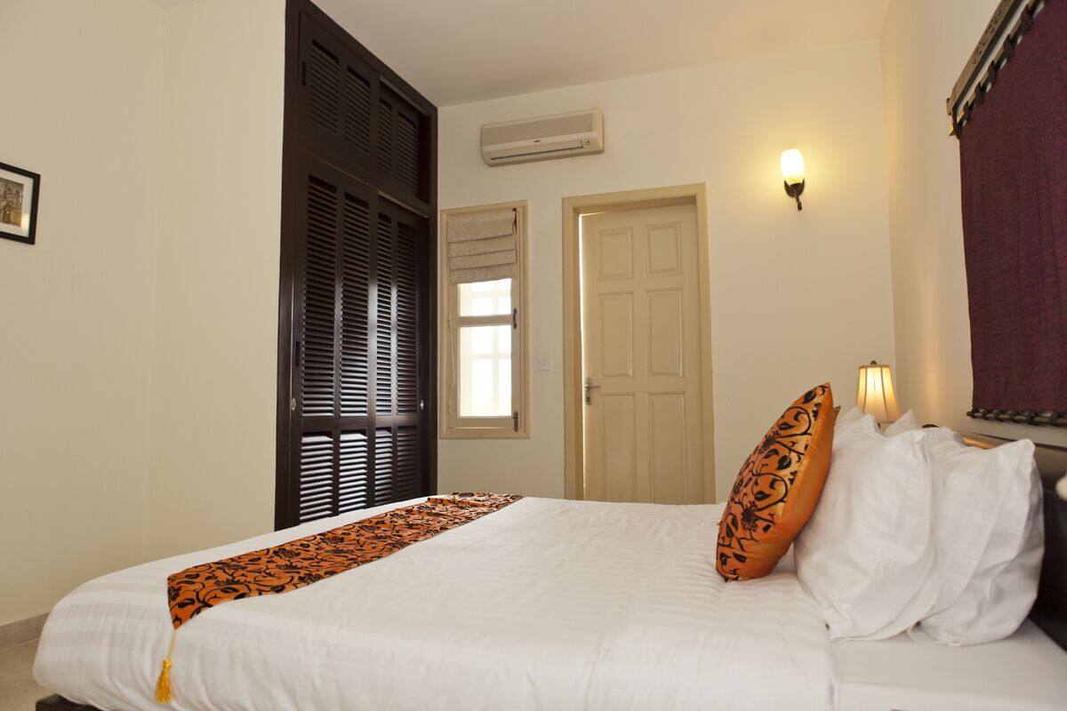 A neatly made bed with a patterned bedspread, a closed door, and a wall-mounted light fixture.
