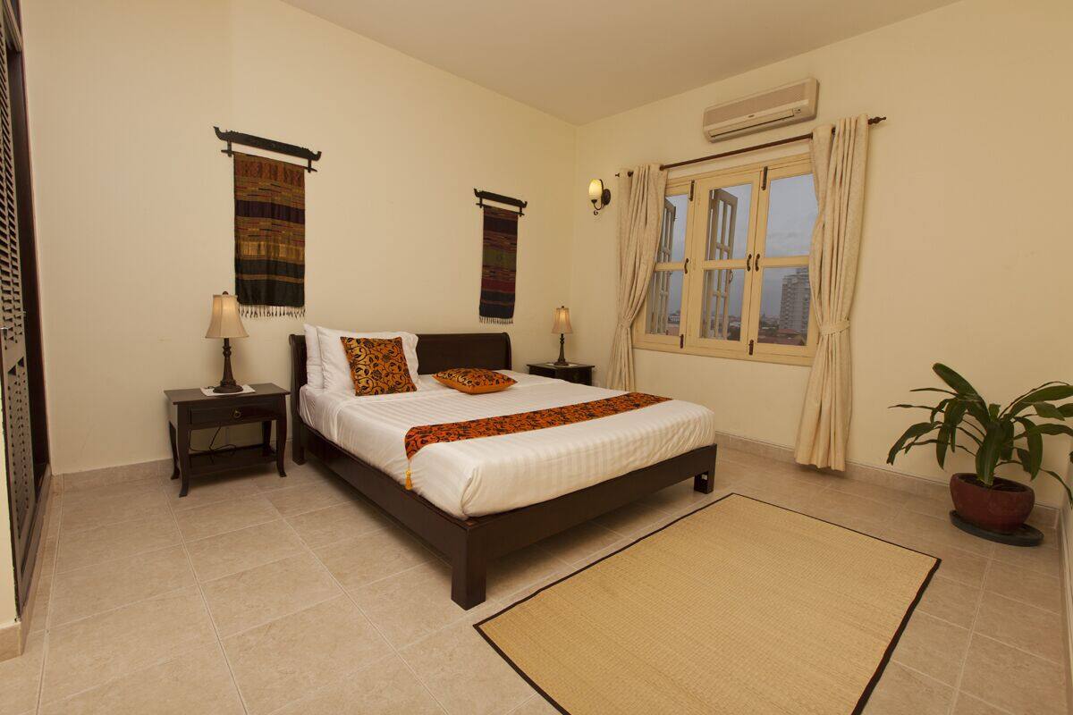 A bedroom with a bed, two nightstands, a window with curtains, a wall-mounted air conditioner, and a potted plant.