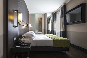 A hotel room with a bed, a desk with a telephone, a TV mounted on the wall, and a mirror.