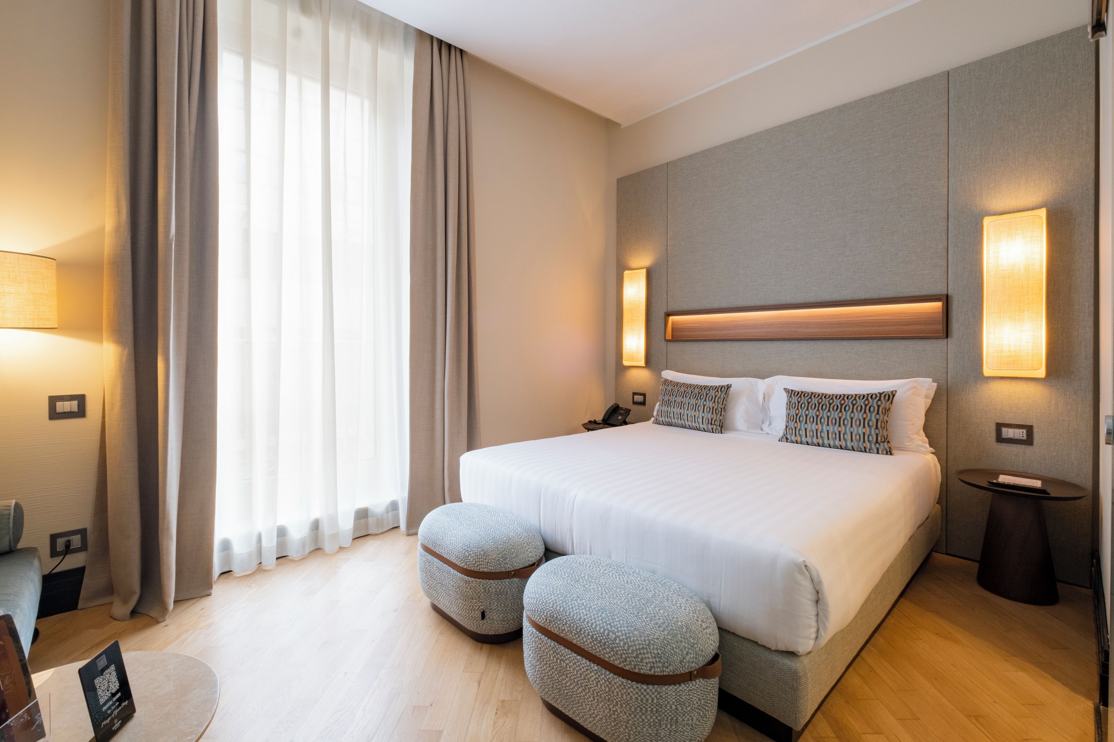 A hotel room with a large bed, two ottomans, a bedside table, a lamp, and a view of the outdoors.