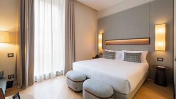 A hotel room with a large bed, two ottomans, a bedside table, a lamp, and a view of the outdoors.