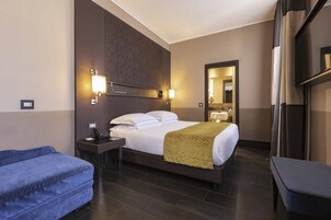 A hotel room with a large bed, a desk with a telephone, a blue sofa, and a bench.