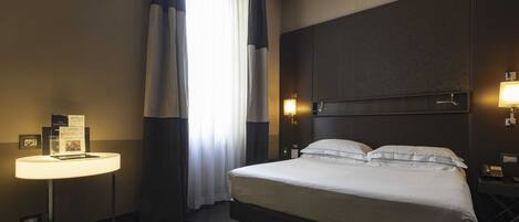 A hotel room with a bed, bedside table, lamp, and a view of the outdoors through the window.