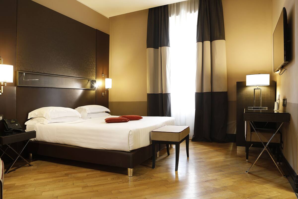 A hotel room with a large bed, bedside table, and a desk with a lamp.