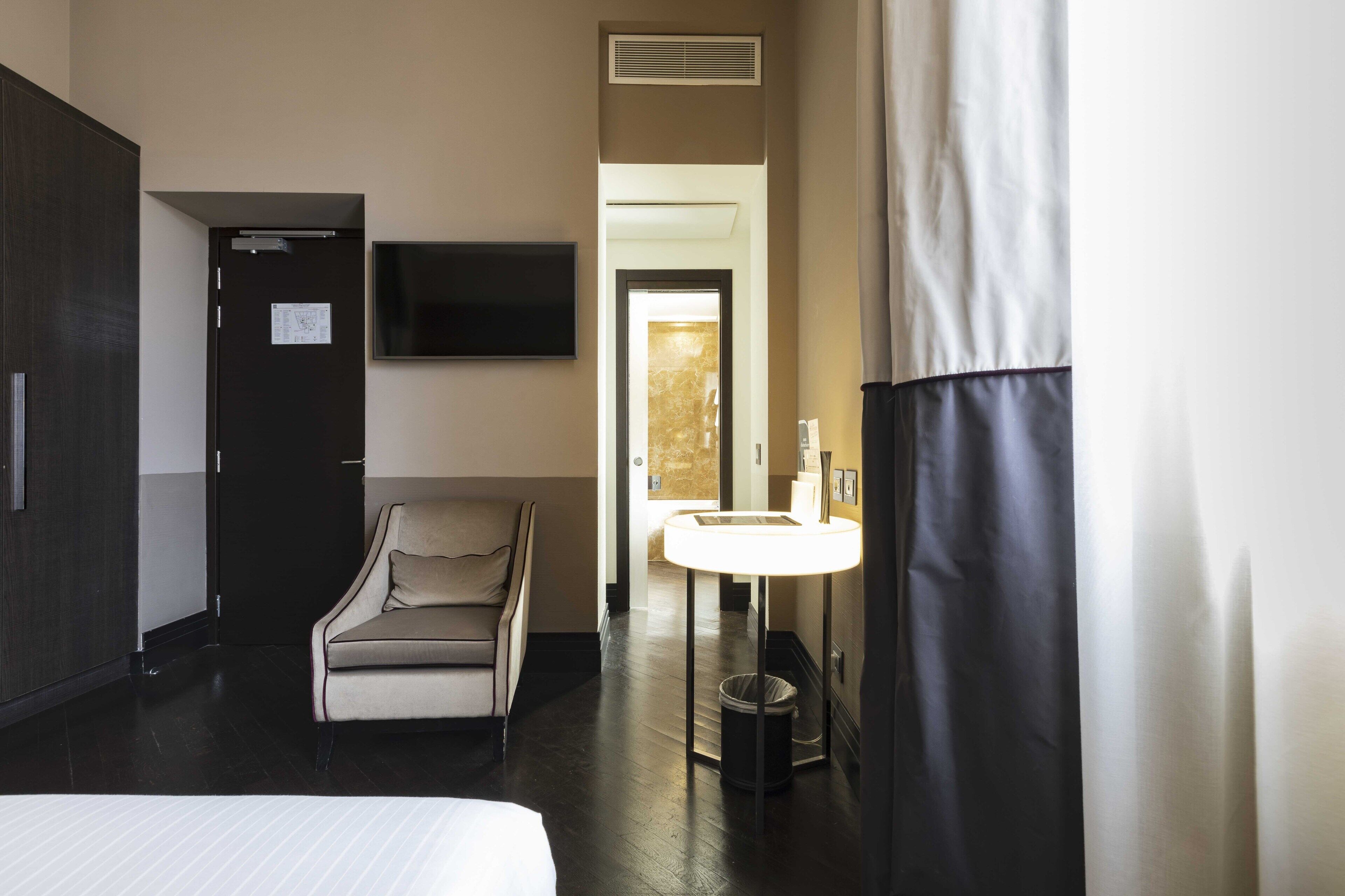 A hotel room with a bed, a sofa, a desk with a lamp, a TV, and a bathroom with a sink and mirror.