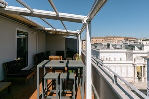 En balkon med et bord, stole og en sofa, samt udsigt til bygninger i baggrunden.