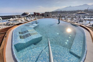 Une grande piscine à débordement, aux eaux cristallines, offrant une vue sur le port de plaisance et les montagnes en arrière-plan.
