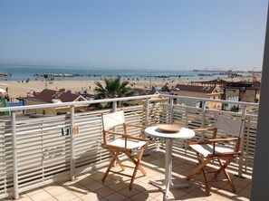 A balcony with two wooden chairs and a small round table, overlooking a beach with people and boats.