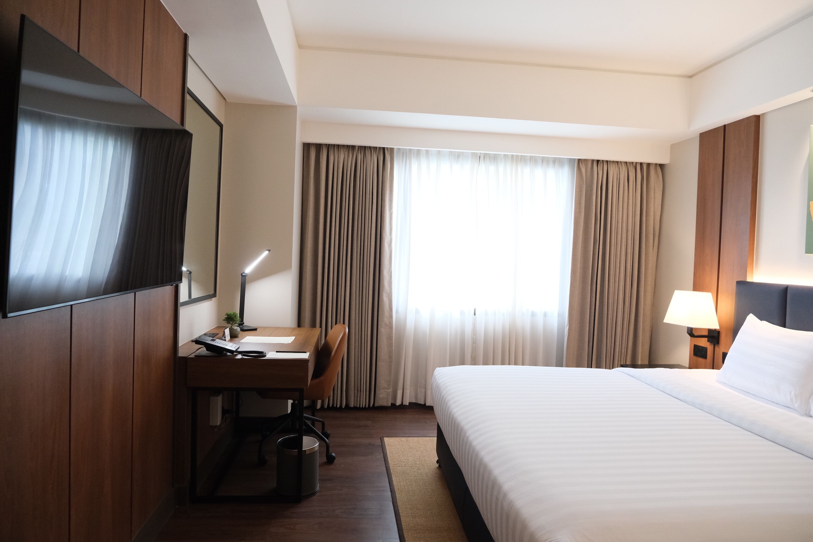A hotel room with a bed, desk, chair, lamp, and curtains.