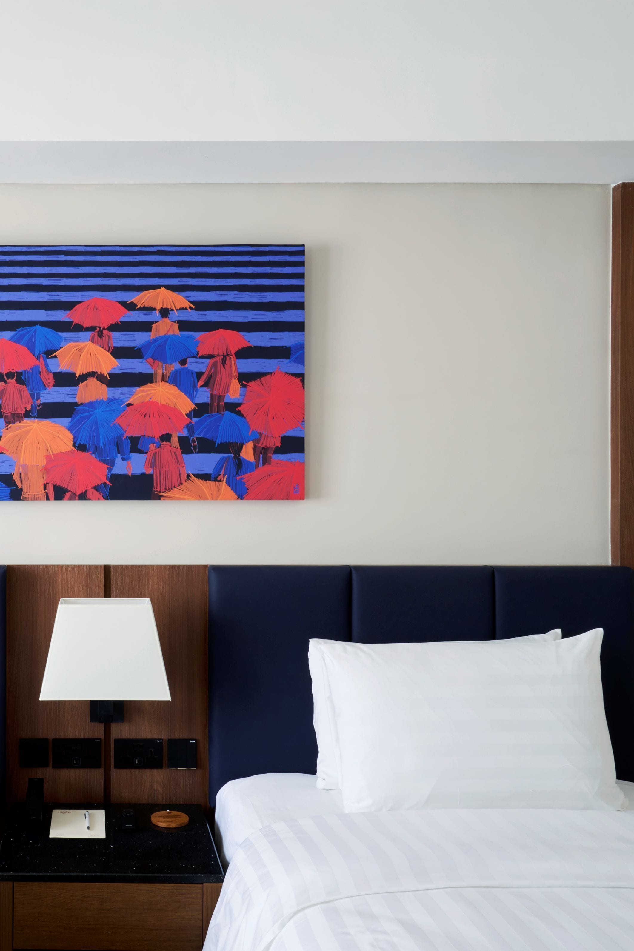 A hotel room with a bed, a nightstand with a lamp, and a wall-mounted painting of umbrellas.
