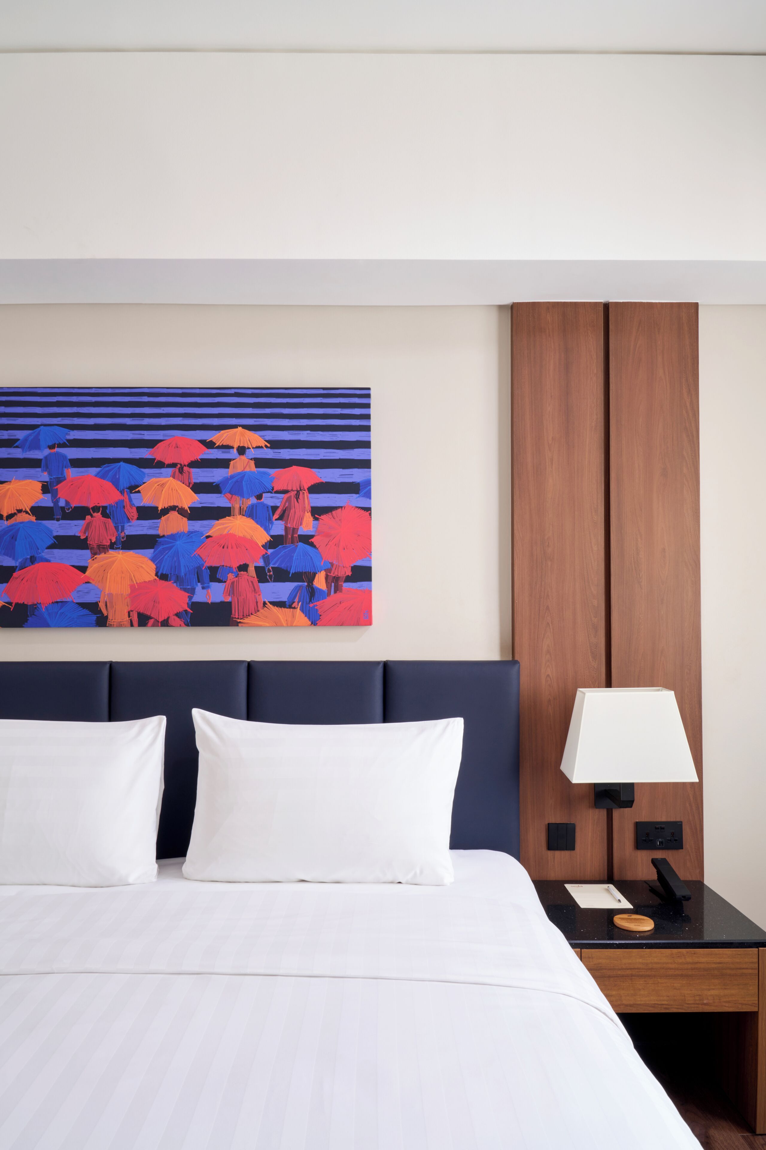 A hotel room with a bed, a nightstand with a lamp, a wooden panel wall, and a colorful painting above the bed.
