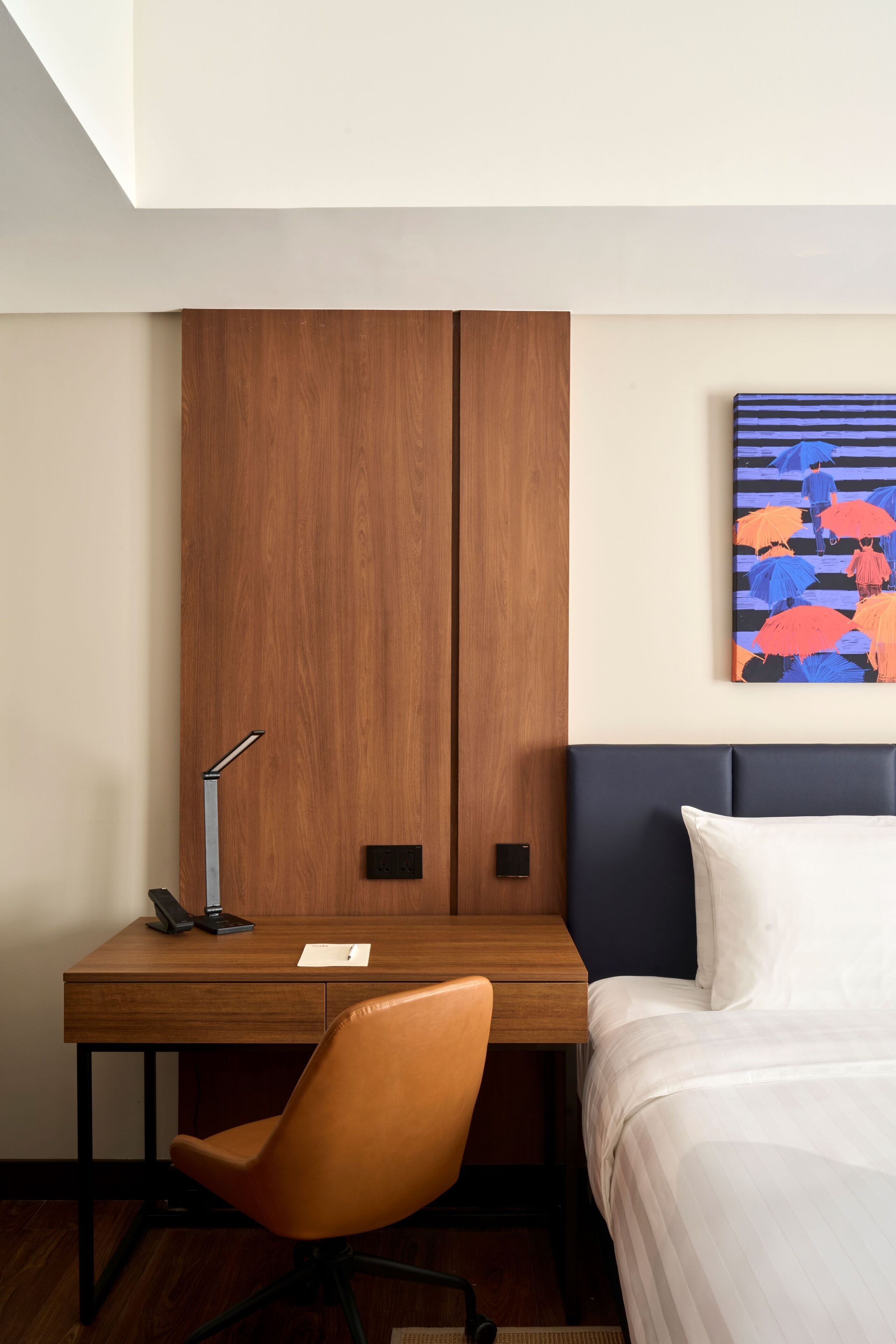 A hotel room with a wooden desk, a chair, a bed, and a painting on the wall.