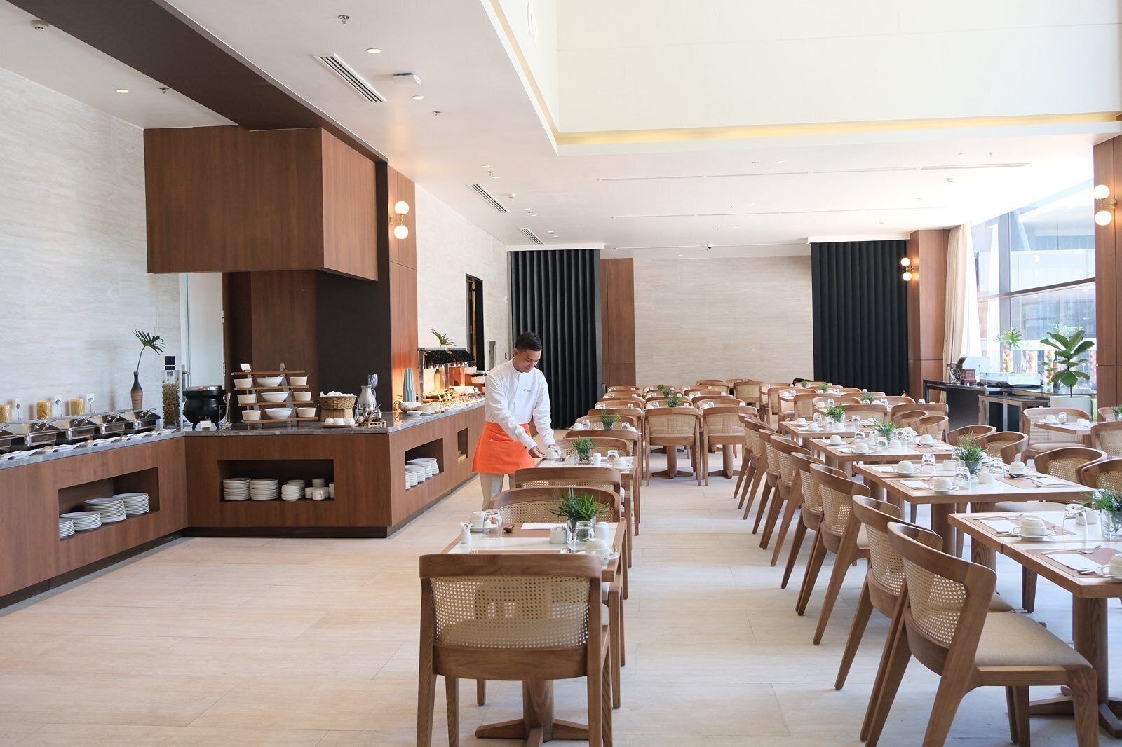 Uma ampla área de refeições com longas mesas e cadeiras de madeira, um balcão de buffet com diversos pratos, e um design de interiores moderno.
