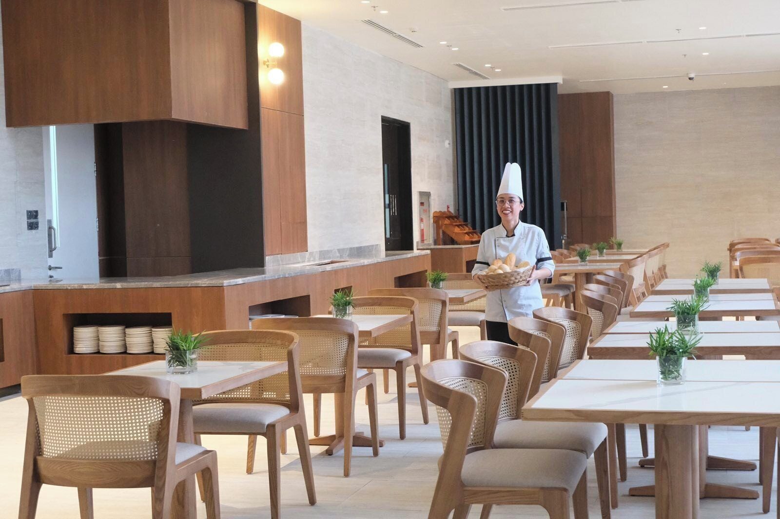 A modern dining area with wooden tables and chairs, a chef serving bread, and a kitchen area with wooden cabinets and a large screen.